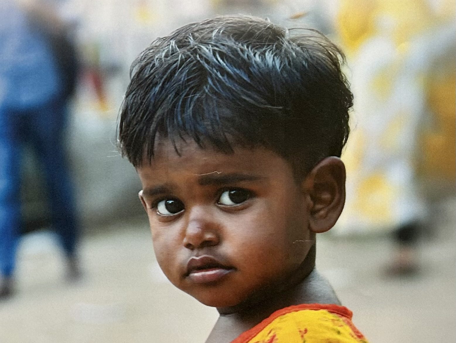 Face of a young child from South Asia wearing a red and yellow shirt.
